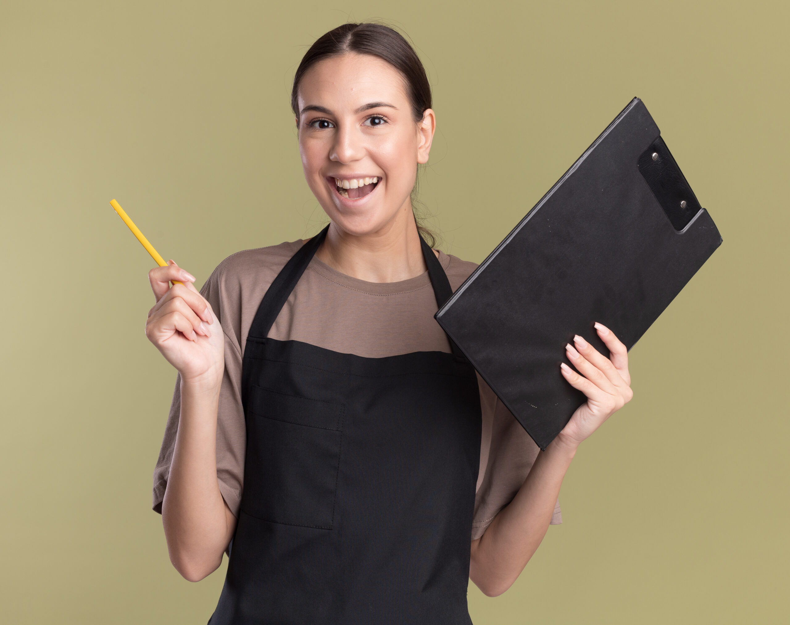 joyful young brunette barber girl in uniform holds pencil and clipboard isolated on olive green background with copy space